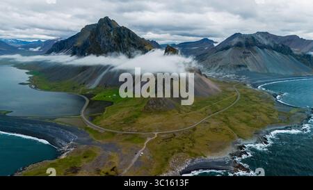 Des routes sinueuses traversent les champs verdoyants menant aux montagnes imposantes le long de la côte islandaise. Des nuages spectaculaires enveloppent des sommets, créant un panorama à couper le souffle pendant une journée tranquille. Banque D'Images