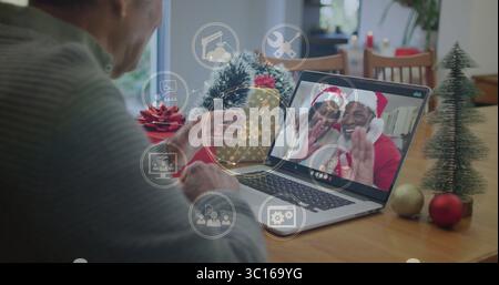 Homme senior dans le pull agitant à l'ordinateur portable pendant l'appel à la table à manger, avec des icônes d'interface Banque D'Images