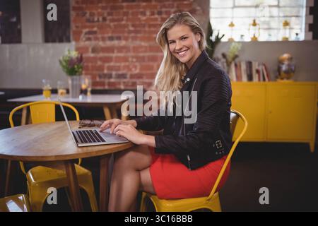 Femme tapant sur un ordinateur portable argenté à la table ronde dans un café avec des chaises jaunes et un mur de briques Banque D'Images