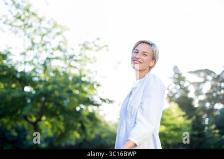 Femme au début des années 30 debout dans un parc ensoleillé parmi le feuillage portant une robe légère, espace de copie Banque D'Images
