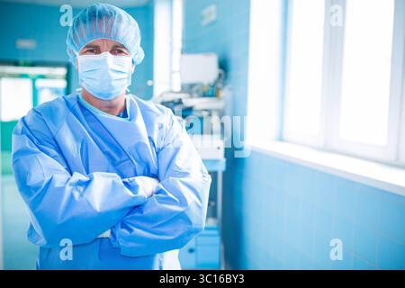 Chirurgien masculin debout dans le couloir chirurgical carrelé bleu à côté du chariot d'équipement sous des fenêtres lumineuses Banque D'Images
