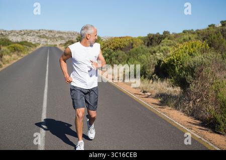 Coureur masculin senior jogging sur une route asphaltée droite avec une ligne centrale blanche sous ciel dégagé Banque D'Images