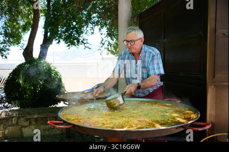 Scène de cuisine en plein air avec un chef préparant une paella dans une fête de village espagnol. Une grande casserole à paella et une ambiance festive sont les points forts Banque D'Images
