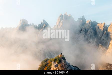 Vue aérienne de pics dentelés perçant à travers des nuages éthérés, une figure solitaire debout au sommet d'un éperon rocheux, Parco Naturale Tre Cime, Vénétie, Italie. Banque D'Images