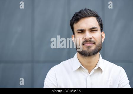 Un bel homme d'origine indienne se tient confiant, posant devant un bâtiment moderne, texturé et gris. Banque D'Images