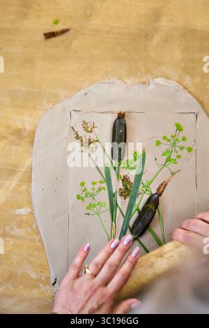 Presser les fleurs et les feuilles en argile avec un rouleau à pâtisserie pour créer une empreinte botanique pour le relief du plâtre. Mains aplatissant les plantes dans le bricolage étape par étape t Banque D'Images
