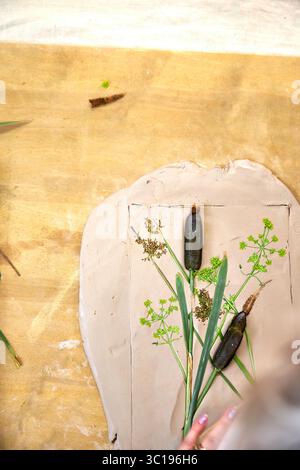 Presser les fleurs et les feuilles en argile avec un rouleau à pâtisserie pour créer une empreinte botanique pour le relief du plâtre. Mains aplatissant les plantes dans le bricolage étape par étape t Banque D'Images