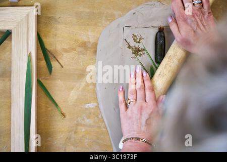 Presser les fleurs et les feuilles en argile avec un rouleau à pâtisserie pour créer une empreinte botanique pour le relief du plâtre. Mains aplatissant les plantes dans le bricolage étape par étape t Banque D'Images