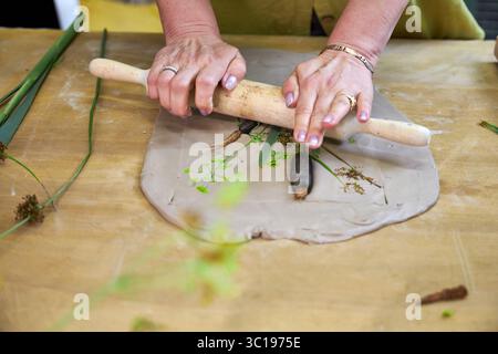 Presser les fleurs et les feuilles en argile avec un rouleau à pâtisserie pour créer une empreinte botanique pour le relief du plâtre. Mains aplatissant les plantes dans le bricolage étape par étape t Banque D'Images