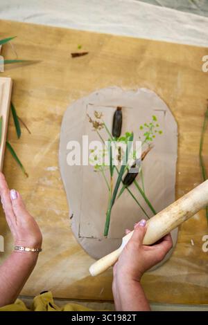 Presser les fleurs et les feuilles en argile avec un rouleau à pâtisserie pour créer une empreinte botanique pour le relief du plâtre. Mains aplatissant les plantes dans le bricolage étape par étape t Banque D'Images