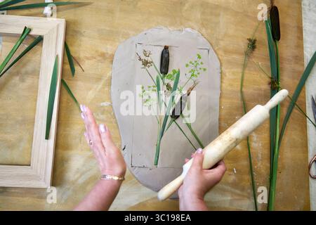 Presser les fleurs et les feuilles en argile avec un rouleau à pâtisserie pour créer une empreinte botanique pour le relief du plâtre. Mains aplatissant les plantes dans le bricolage étape par étape t Banque D'Images