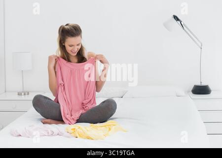Femme tenant le haut rose tout en comparant les chemises rose pâle et jaune sur le lit dans la chambre minimaliste Banque D'Images