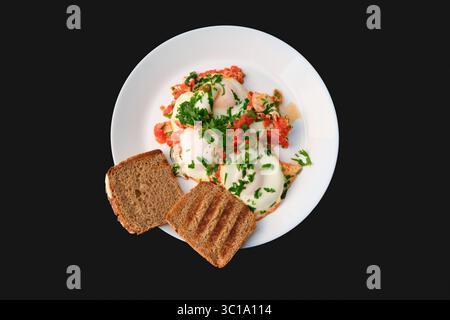 Shakshuka avec des œufs et du persil servi avec du pain grillé sur une assiette blanche, plat sur fond noir, concept de petit déjeuner végétarien sain. Banque D'Images