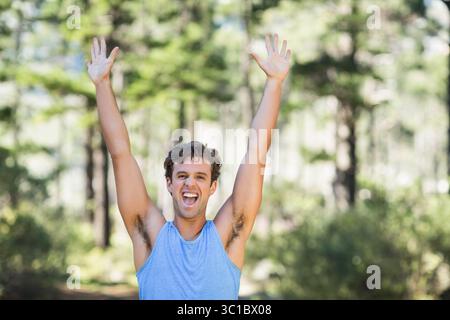 Homme à la fin de la vingtaine levant les bras au-dessus de la tête dans la forêt ensoleillée de défrichement portant une chemise bleue sans manches Banque D'Images