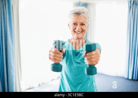 Femme senior soulevant des haltères sarcelle et portant une chemise d'exercice sarcelle dans la zone d'exercice à la maison Banque D'Images