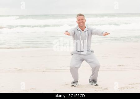 Homme mûr accroupi avec les bras étendus portant un sweat à capuche gris clair et un pantalon de survêtement sur le rivage de sable Banque D'Images
