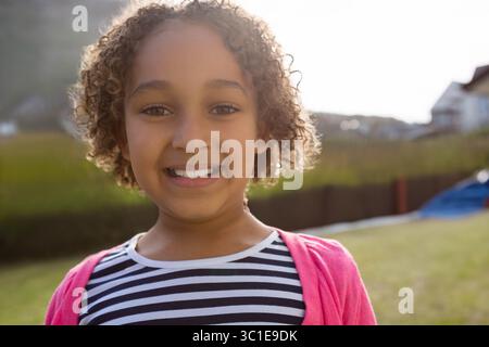 Enfant fille debout dans la cour herbeuse portant chemise rayée et cardigan rose sous le ciel lumineux Banque D'Images