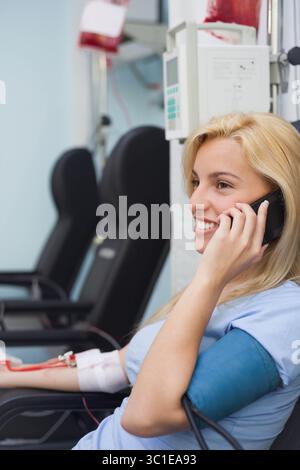 Donneur de sang féminin assis dans le fauteuil de la clinique parlant sur smartphone connecté à la ligne IV, espace de copie Banque D'Images
