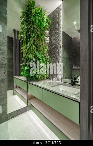 Meuble-lavabo moderne avec armoire verte, plateau en marbre gris et jardin vertical luxuriant. Design d'intérieur biophilique avec carreaux hexagonaux et miroir. Banque D'Images
