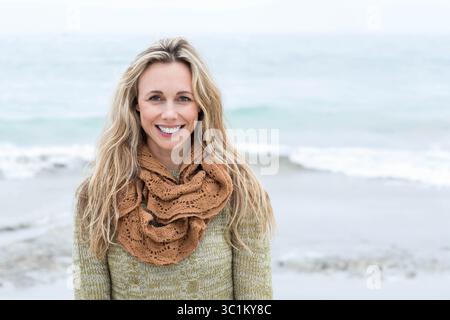 Femme debout sur la plage souriant à la caméra portant un pull vert olive avec une écharpe brune texturée Banque D'Images