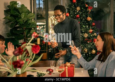Un jeune couple diversifié célébrant les fêtes, riant de joie alors que l'homme ouvre une bouteille de champagne à côté d'un sapin de Noël lors d'un ho festif Banque D'Images