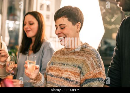 Une belle jeune femme aux cheveux courts éclate de rire authentique tout en tenant un verre de vin mousseux lors d'une fête animée avec des amis. Banque D'Images