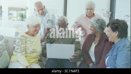Rassemblement de personnes âgées portant des vêtements décontractés collaborant autour d'un ordinateur portable argenté dans un salon résidentiel Banque D'Images