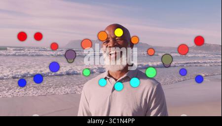 Homme senior debout portant un polo gris clair sur le rivage de l'océan, avec recouvrement de cercles arc-en-ciel Banque D'Images
