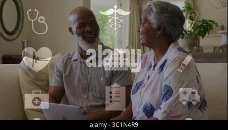 Couple mûr souriant discutant d'informations de santé sur ordinateur portable dans le canapé de la maison, avec des icônes médicales flottantes Banque D'Images