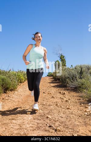 Femme adulte moyenne qui fait du jogging sur une piste de saleté rougeâtre au milieu de broussailles portant des chaussures de course blanches Banque D'Images
