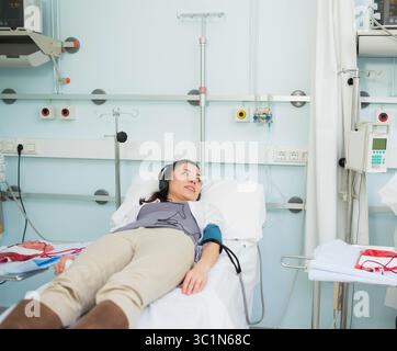Inclinaison sur le lit d'hôpital, port d'écouteurs et brassard de tensiométrie près de la tige porte-perfusion avec tubulure Banque D'Images