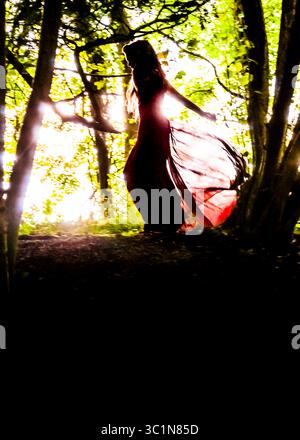 Une femme dans une robe rouge fluide se silhouette contre la lumière du soleil brillante filtrant à travers les arbres. Banque D'Images