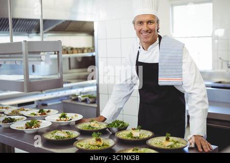 Chef masculin portant uniforme préparant des plats à table sous des lampes chauffantes dans une cuisine occupée, espace de copie Banque D'Images