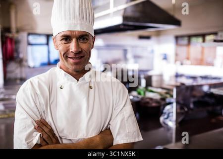 Chef mûr debout dans la cuisine du restaurant portant uniforme et toque avec comptoirs en acier inoxydable Banque D'Images
