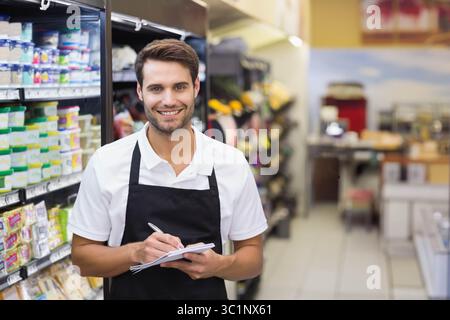 Employé de l'épicerie portant un tablier noir au-dessus d'une chemise blanche tenant un stylo et un bloc-notes dans l'allée de l'épicerie Banque D'Images