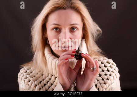 Une femme à la peau claire aux cheveux blonds et aux yeux bleus tient une pomme de pin dans ses mains. Elle porte un pull en tricot texturé de couleur crème et un s. Banque D'Images