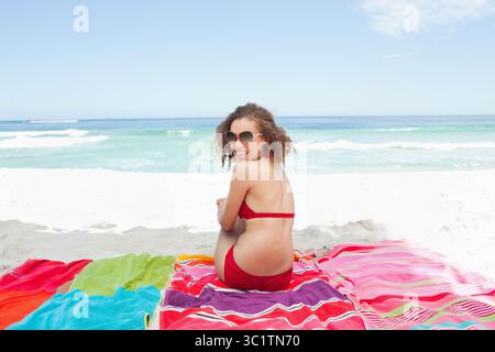 Femme enceinte assise sur des serviettes de plage rayées sur le rivage portant un bikini rouge et des lunettes de soleil surdimensionnées Banque D'Images