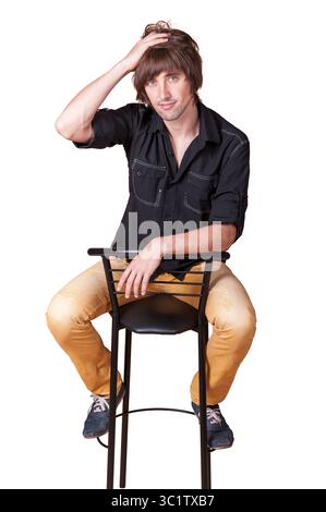 Un jeune homme dans une chemise noire et un pantalon bronzé est assis sur un tabouret, contemplant de manière créative. Banque D'Images