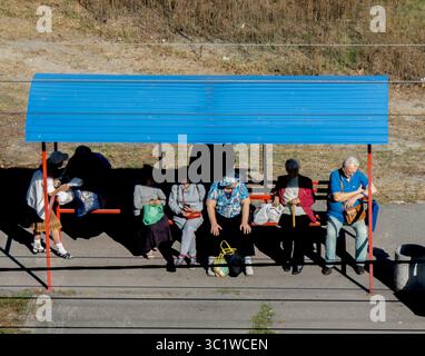 Kiev, Ukraine - 20 juillet 2025 : groupe de personnes âgées assis sur un banc sous un toit bleu à la gare rurale, attendant le train le jour ensoleillé d'automne Banque D'Images