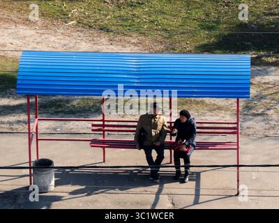 Kiev, Ukraine - 20 juillet 2025 : deux personnes âgées assises sur un banc sous un arrêt de bus avec un toit en tôle ondulée bleue, parlant en attendant les transports en commun Banque D'Images