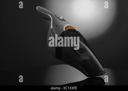 Un aspirateur à main élégant, noir et gris, est mis en évidence sur un fond sombre et réfléchissant. Une source lumineuse circulaire brillante brille Banque D'Images