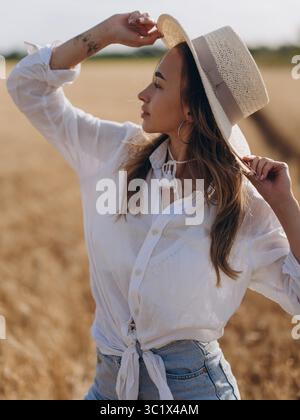 Jeune femme en chapeau de paille, chemise blanche et short en denim se tient dans le champ de blé doré et jouit par la vue. Scène estivale rurale paisible avec beau naturel Banque D'Images