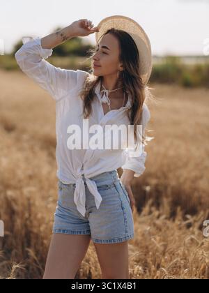 Jeune femme en chapeau de paille, chemise blanche et short en denim se tient dans le champ de blé doré et jouit par la vue. Scène estivale rurale paisible avec beau naturel Banque D'Images