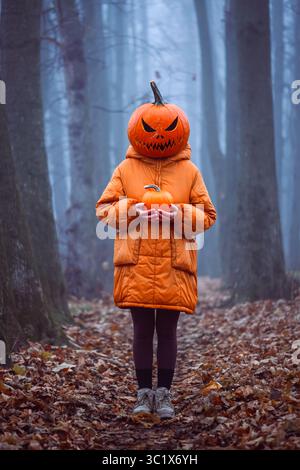 Fille d'Halloween dans le masque de citrouille sculpté se dresse dans la forêt d'automne brumeuse avec une petite citrouille dans les mains. Moment effrayant dans les bois rempli de mystère, de costumes et d'humeur automnale Banque D'Images