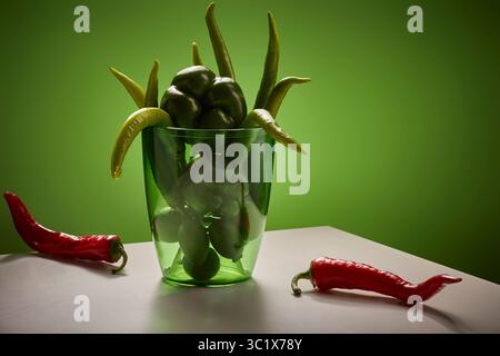 Un vase en verre rempli de piments verts repose sur une table blanche. Un seul piment rouge repose sur la table à côté du vase. L'arrière-plan est un Banque D'Images