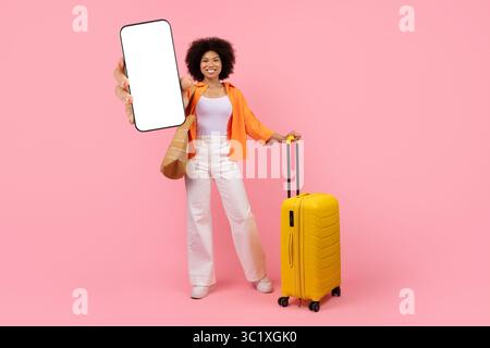 Souriante jeune femme afro-américaine tenant une grande maquette de téléphone et une valise de voyage Banque D'Images