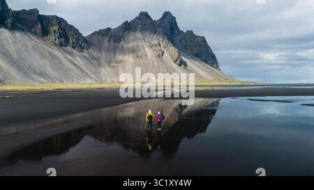 Deux aventuriers se tiennent sur la plage de sable noir sereine de Stokksnes, en Islande, contemplant la majestueuse montagne Vestrahorn et capturant les reflets dans des eaux calmes peu profondes au cours d'une journée paisible. Banque D'Images