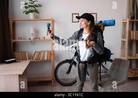 Portrait de jeune femme randonneur enregistrant la vidéo sur la caméra d'action debout avec sac à dos et équipement de randonnée près de la table à la maison. Copier l'espace Banque D'Images