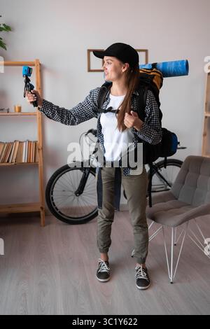 Portrait de jeune femme randonneur enregistrant la vidéo sur la caméra d'action debout avec sac à dos et équipement de randonnée près de la table à la maison. Copier l'espace Banque D'Images