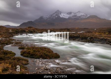 Vue d'une rivière qui coule rapidement en cascade à travers un paysage rocheux accidenté sous un ciel couvant, avec des montagnes enneigées qui se profilent au loin, Écosse, Royaume-Uni. Banque D'Images